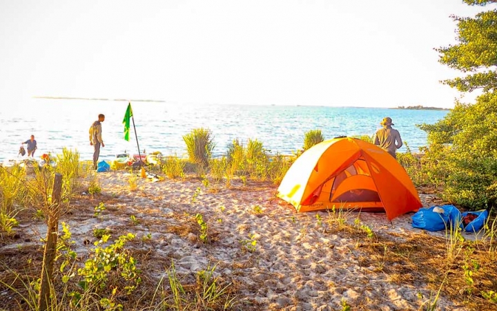 An orange tent rests on the sand of a beach. There are a few people around, and vast blue ocean in the background. 